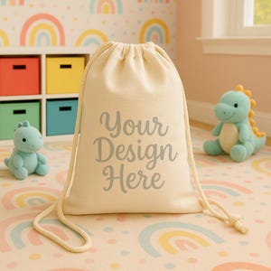 Puede incluir: Una bolsa con cordón de color crema con el texto "Your Design Here" en gris. La bolsa está sobre una alfombra con estampado de arcoíris, con dos juguetes de dinosaurio de peluche y una unidad de almacenamiento colorida en el fondo.