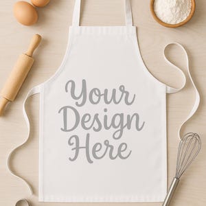 May include: White apron with the text "Your Design Here" in gray script. The apron is surrounded by baking tools and ingredients, including eggs, flour, a rolling pin, a whisk, and a cookie cutter. The apron is a blank canvas for custom designs.