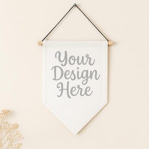 May include: White pennant banner with the text "Your Design Here" in gray script. The banner is suspended from a wooden dowel and black string, mounted on a light beige wall. Dried flowers are in the lower left corner.