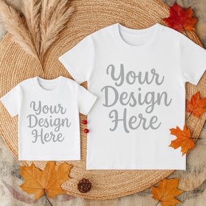 May include: Two white t-shirts, one adult and one child size, with the text "Your Design Here" in gray script. The shirts are on a woven mat with fall foliage, including red and orange leaves, and a pinecone.