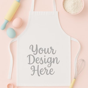 May include: A white apron with the text "Your Design Here" is surrounded by baking tools and ingredients. A light blue and yellow rolling pin, two eggs, a bowl of flour, a whisk, and a cookie cutter are arranged on a pink background.