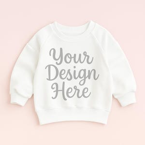 以下が含まれることがあります： グレーのスクリプトで「Your Design Here」というテキストが入った白いスウェットシャツ。スウェットシャツはクルーネックと長袖です。背景は薄いピンク色です。