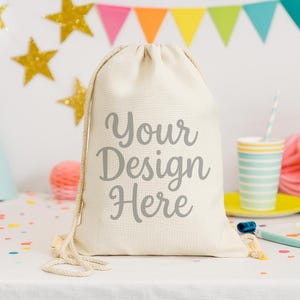 Puede incluir: Una bolsa con cordón de color crema con el texto "Your Design Here" en gris. La bolsa está sobre una superficie blanca con decoraciones de fiesta en el fondo, incluyendo estrellas doradas y una pancarta colorida.