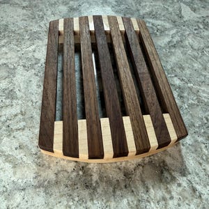 May include: A wooden trivet with a rectangular shape and a curved edge. The trivet is made of alternating dark brown and light tan wooden slats. The slats are arranged in a parallel pattern, creating a visually appealing design.