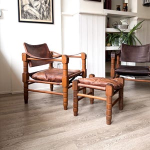 May include: Two armchairs and a matching ottoman featuring light brown wooden frames with turned legs and dark brown leather sling-style seats and backrests. The leather cushions are stitched with a grid pattern and secured to the frames with straps.