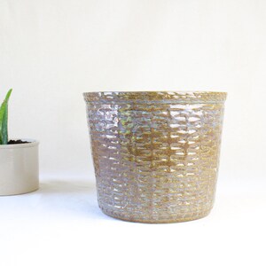 May include: A ceramic plant pot with a textured, grid-like pattern in shades of brown and green. The pot is cylindrical and sits on a white surface. A smaller white pot with a green plant is to the left.