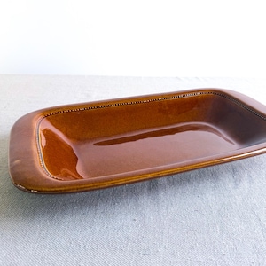 May include: A rectangular, brown ceramic serving dish with a glossy finish. The dish has rounded corners and a beaded border. The interior is smooth and reflective, suitable for serving food.