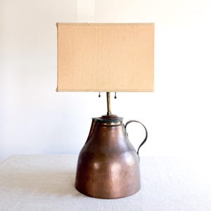 May include: A table lamp featuring a base made from a large, rounded copper pitcher with a curved handle and a rectangular, beige fabric lampshade. The lamp includes a central brass-colored rod and two small pull chains hanging beneath the shade.