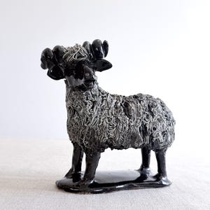 May include: A ceramic ram figurine featuring a glossy black finish on the head, legs, and base, contrasted by a textured, shaggy grey coat. The ram has prominent curled horns and stands on an oval-shaped base, showcasing detailed sculptural work.