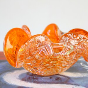 May include: An orange glass bowl with a unique, organic shape. The bowl has a textured surface with iridescent accents. The bowl is approximately 6 inches in diameter and is likely used for decorative purposes.
