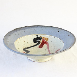 May include: A ceramic bowl with a light blue rim and a cream-colored interior. The interior features abstract black, red, and gold brushstrokes. The bowl has a small, white pedestal base.