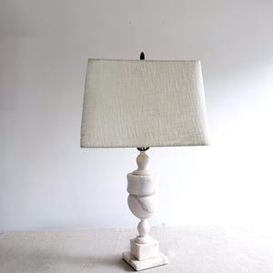 May include: A white marble table lamp with a rectangular lampshade. The lamp base has a stacked, urn-like design. The lampshade is a light beige color. The lamp is on a light-colored surface.