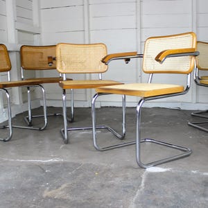 May include: A set of five vintage chairs with chrome frames, wooden seats, and woven cane backs. One chair has armrests. The chairs are arranged in a row against a white wall and gray floor.