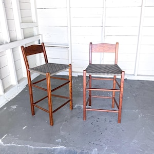 May include: Two wooden bar stools with woven seats. The stools have a dark brown finish and a woven seat in shades of gray and black. The stools are positioned against a white wall.