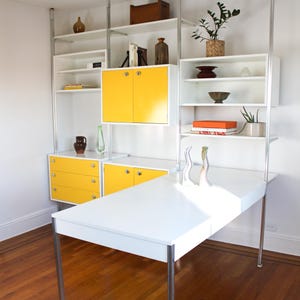 May include: A modular wall unit featuring white shelves, yellow cabinets, and a white desk surface supported by silver metal poles. The unit includes a three-drawer chest, open shelving for decor, and a desk extension, all mounted on a vertical frame.