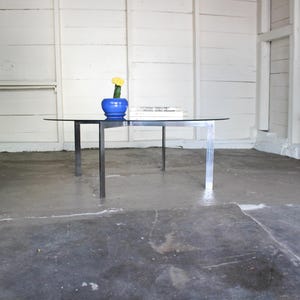 May include: A modern glass-top coffee table with a black metal frame and legs. A blue ceramic pot with a yellow flower and a stack of books are on the table. The table is in a room with white walls.