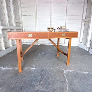 May include: A rectangular wooden desk featuring two drawers with brass-colored recessed pulls. The desk is supported by four straight legs connected by diagonal cross-bracing supports. Two small decorative bowls sit on the flat top surface.