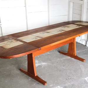May include: A wooden dining table with a dark brown tabletop and light brown legs. The tabletop features decorative tiles with a light beige background and a dark brown design. The table is approximately 72 inches long.