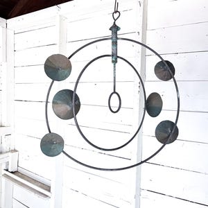 May include: A hanging metal sculpture with a series of concentric circles and circular, disc-shaped accents. The metal has a dark, aged appearance with hints of green. The sculpture is suspended from a hook and ring.