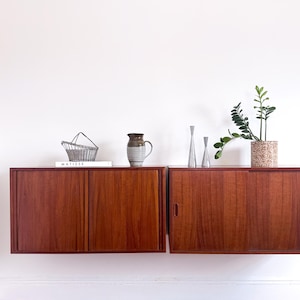 May include: Two wall-mounted, mid-century modern style cabinets with sliding doors. The cabinets are made of reddish-brown wood. Decorative items on top include a wire basket, a ceramic pitcher, and a potted plant.