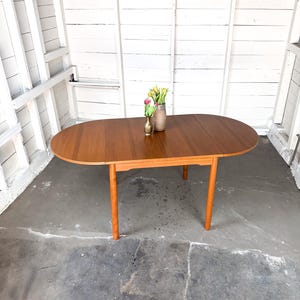 May include: An oval wooden dining table with a warm brown finish and four tapered legs. The tabletop features a visible wood grain pattern and a central seam. A small brass vase containing yellow and pink tulips sits in the center of the table.