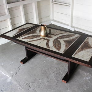 May include: A rectangular dark brown wooden table with a decorative tile top. The tile has a beige background with brown leaf-like patterns. A small, gold-colored pitcher sits on the table. The table is approximately 60 inches long.