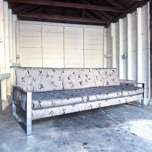 May include: A three-seat sofa featuring a polished chrome metal frame and light gray fabric upholstery with a dark gray and black floral pattern. The sofa has a rectangular backrest, a long seat cushion, and two armrests with a metallic finish.