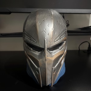 May include: A silver-colored full-face mask featuring a textured, battle-worn finish with numerous small indentations across the surface. The mask has a central vertical ridge, narrow horizontal eye openings, and a pointed chin section.