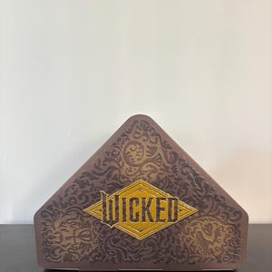 Puede incluir: Una caja triangular marrón con patrones florales en relieve. La palabra "WICKED" se muestra en letras doradas dentro de un rombo dorado en la parte delantera de la caja.