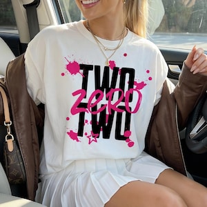 May include: A white t-shirt with the text "TWO ZERO TWO" in black and pink, with pink paint splatters. The shirt is worn with a white pleated skirt and a brown jacket. The person is sitting in a car.