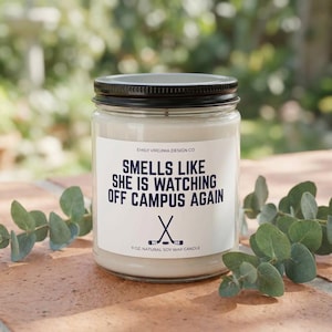 Puede incluir: Vela en tarro de vidrio transparente con tapa negra. La etiqueta dice "SMELLS LIKE SHE IS WATCHING OFF CAMPUS AGAIN" con palos de hockey. La vela es de 255g y está hecha de cera de soja natural.