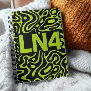 May include: A spiral-bound notebook with a black cover featuring a neon green abstract design and the letters "LN4". The notebook is resting on a white knitted blanket.