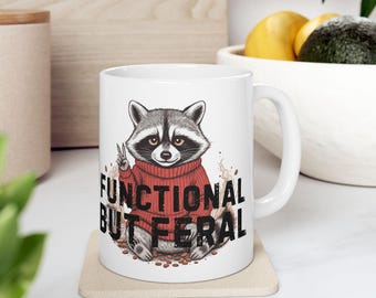 May include: A white ceramic mug featuring a graphic illustration of a raccoon wearing a red jumper and making a peace sign. The text "FUNCTIONAL BUT FERAL" is printed in bold black capital letters below the raccoon, surrounded by small coffee bean icons.