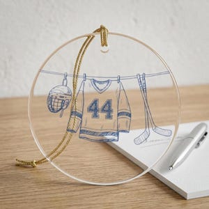May include: A circular clear acrylic ornament featuring a blue line drawing of a hockey helmet, a jersey with the number 44, and two hockey sticks hanging on a clothesline. The ornament includes a gold hanging cord.