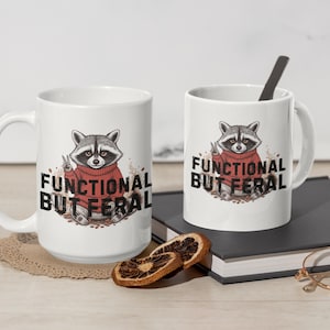 May include: Two white ceramic mugs with a raccoon graphic wearing a red sweater and the text "FUNCTIONAL BUT FERAL". One mug is larger than the other. A black spoon rests in the smaller mug.