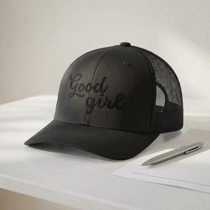 May include: Black baseball cap with the embroidered text "Good girl". The cap features a mesh back and a curved brim. A silver pen and a piece of white paper are next to the cap.