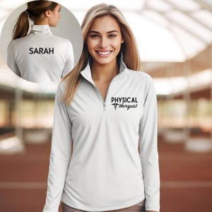 May include: White long-sleeve athletic shirt with a quarter-zip collar. The shirt features the text "PHYSICAL therapist" in black on the front and the name "SARAH" on the back. The shirt is designed for comfort and performance.