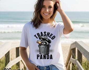 May include: White t-shirt with a graphic of a raccoon in a trash can, holding a bottle. The text "Trashed Panda" is above and below the graphic. The background is a beach scene.