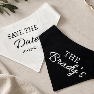 May include: Two triangular pet bandanas, one white and one black. The white bandana features black text reading SAVE THE Date 10.23.27. The black bandana features white embroidered text reading THE Brady's. Both items are made of fabric.