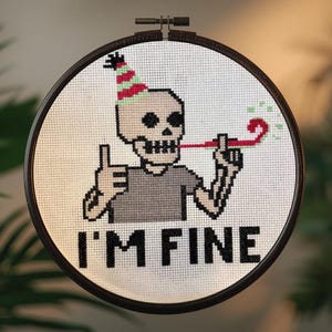 May include: Cross-stitch artwork in a round frame, depicting a skeleton wearing a party hat and blowing a party horn. The text "I'M FINE" is stitched below. The frame is dark wood.