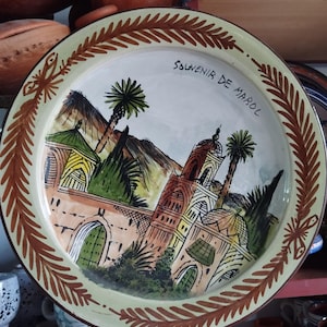 Peut inclure: Assiette en céramique peinte à la main avec une illustration détaillée d'un bâtiment, de palmiers et de montagnes. L'assiette a une bordure de feuilles marron et l'inscription "Souvenir de Maroc".