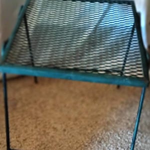 May include: A teal metal folding table featuring a rectangular top with a diamond-patterned mesh surface. The table has thin, straight legs and a frame constructed from slender metal tubing, providing a simple and functional design structure.
