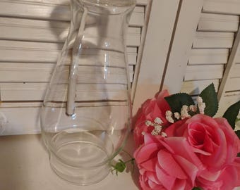 May include: A clear glass water pitcher with a decorative rim and a long, slender shape. A pink floral arrangement with green leaves sits beside the pitcher. The pitcher is empty and appears to be clean.
