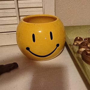 May include: A yellow ceramic smiley face planter. The planter has a round shape with a wide opening and a glossy finish. The face features two black vertical eyes and a curved black smile.
