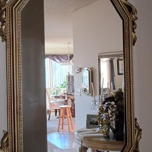 May include: An octagonal wall mirror featuring an ornate, gold-toned frame with intricate scrollwork and a decorative shell-like crest at the top. The reflective glass surface displays a room interior, including a wooden table and a light-colored stool.