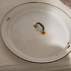 May include: White enamel pot lid with a black rim and a curved, dark handle. The lid shows signs of age with rust spots around the handle. A classic kitchenware item.