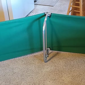May include: A green and silver folding cot. The cot has a rectangular green fabric bed supported by a silver metal frame. The frame has curved legs and a central support. The cot is designed for outdoor or indoor use.