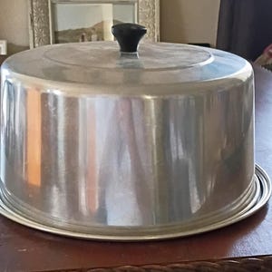 May include: A round, silver-colored metal cake cover with a smooth, reflective surface and a small, black, circular knob handle centered on the top. The cover features a slightly domed top and straight sides that rest on a matching circular base.