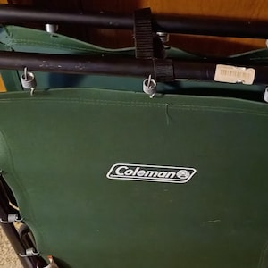 May include: Folded green Coleman camp chair with a black metal frame. The chair has a dark green fabric seat and back, with silver springs and a black strap for securing. The Coleman logo is visible on the front.