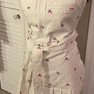 May include: Cream-colored overall dress with a floral pattern in red and black. The dress features a square neckline, pockets, a pleated skirt, and a matching belt. The dress is displayed on a mannequin.